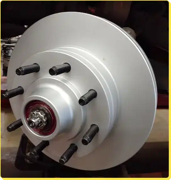 At Jerry's Automotive. Close-up of a shiny, silver disc brake rotor with five lug bolts on a vehicle, showcasing recent brake repair and service. The central hub features a red, capped nut. The rotors smooth, reflective surface indicates its new or recently serviced, contrasting against the darker background components.