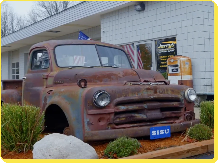 At Jerry's Automotive. A rusty, vintage Dodge pickup truck is displayed outside a building with signs for Jerrys Automotive. The truck has a weathered appearance and is surrounded by shrubs and rocks. An American flag is visible in the background.