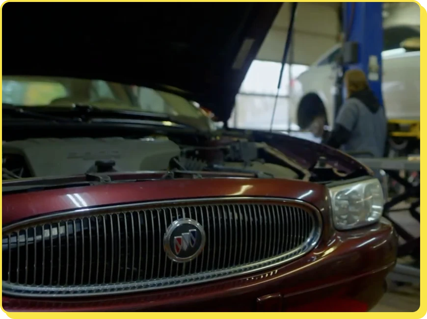 At Jerry's Automotive. A red car with its hood open is inside an auto repair shop. A mechanic in the background is working near another lifted vehicle. The scene suggests maintenance or repair work in progress.