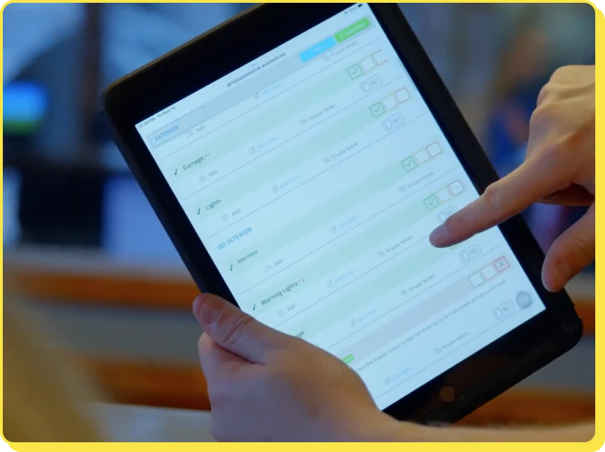 At Jerry's Automotive. A person holds a tablet displaying a checklist or form interface. The screen shows various tasks with checkboxes, some marked completed. The persons fingers interact with the screen. The background is blurred.