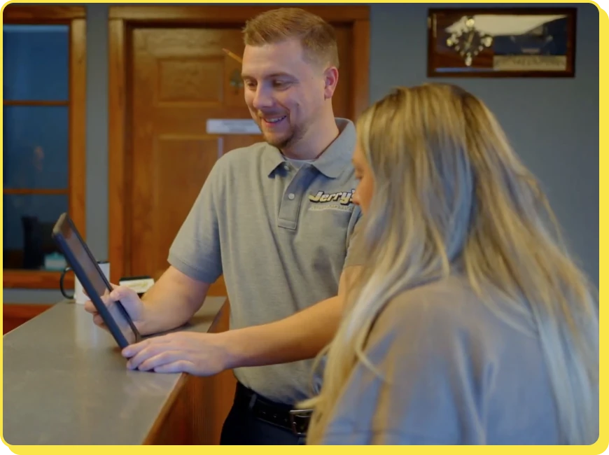 At Jerry's Automotive. A man in a grey polo shirt showing something on a tablet to a woman with long blonde hair at a reception desk. They are in a warmly lit room with wooden doors in the background.