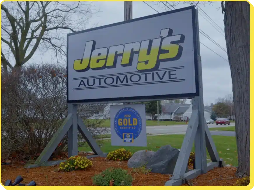 At Jerry's Automotive. A sign for Jerrys Automotive is displayed outdoors. The sign features large, bold letters in yellow and white. Below, a badge reads 2023 Gold Certified. The area is landscaped with flowers, rocks, and grass, with trees and houses in the background.