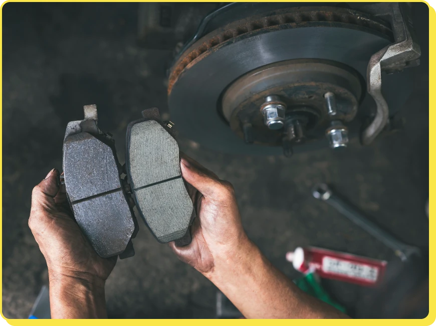 At Jerry's Automotive. Hands holding a new and an old brake pad side by side, showing the difference in thickness. A car brake disc is visible in the background under dim lighting, with a tube of lubricant on the ground nearby.