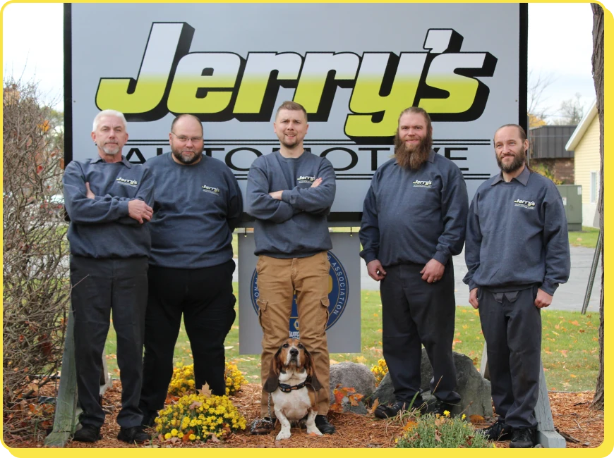 At Jerry's Automotive. Five men wearing gray sweatshirts stand in front of a Jerrys Automotive sign with a dog sitting at their feet. They are outdoors, surrounded by fall foliage and plants.