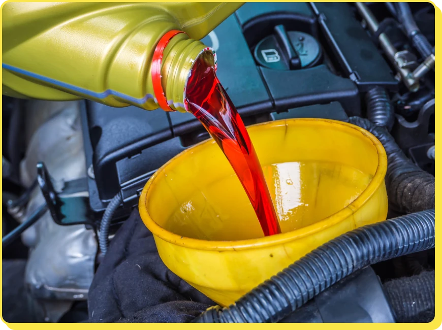 At Jerry's Automotive. A red liquid being poured from a yellow container into a yellow funnel positioned above an engine compartment. Various engine components and cables are visible in the background.