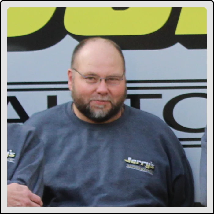 At Jerry's Automotive. A man with a beard and glasses is wearing a gray sweatshirt with Jerrys printed on it. He is standing in front of a vehicle with partially visible text and graphics.