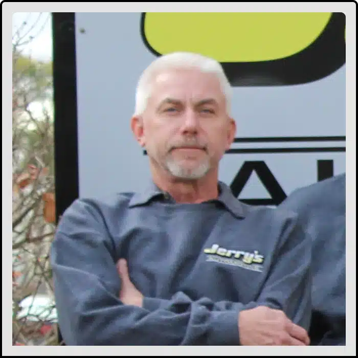 At Jerry's Automotive. A man with short white hair and a beard, wearing a blue-grey sweater with a logo, stands with his arms crossed. A large sign is partially visible behind him, featuring a black and yellow design.