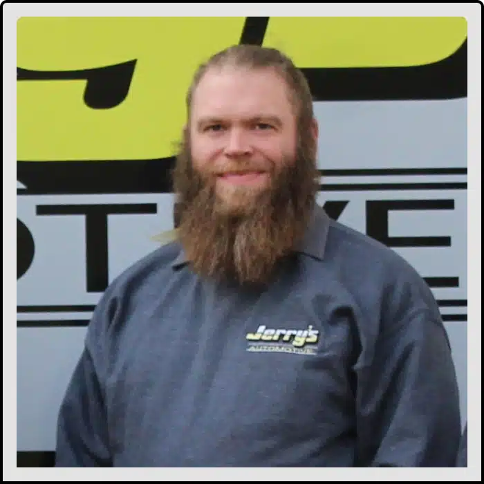 At Jerry's Automotive. A person with a long beard, wearing a dark gray shirt that says Jerrys, stands in front of a sign. The background is partially visible with bold letters.