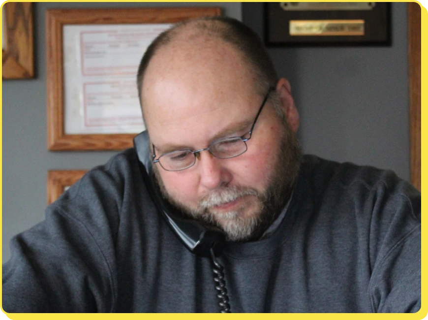 At Jerry's Automotive. A bearded man wearing glasses is sitting and talking on a phone. He looks focused, and there are framed certificates or documents on the wall behind him.